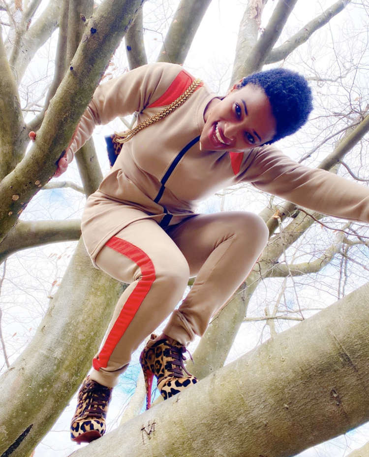 Beautiful black woman dressed in one of Bibi Stewart's designs, a tan sweatsuit. She sits in a tree.